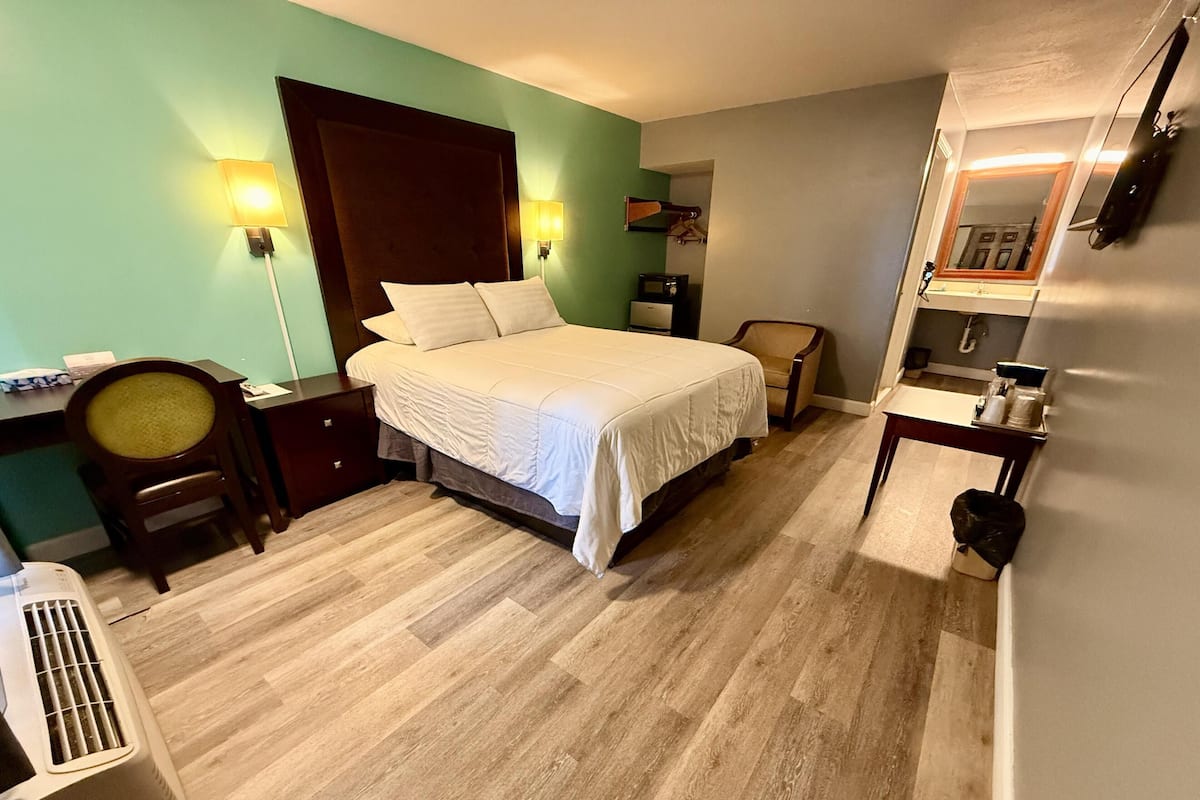 A bedroom with a bed, bedside tables, a chair, a desk, a mirror, and a small kitchenette.