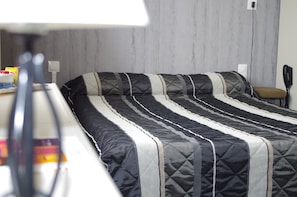 A neatly made bed with a striped quilt, a bedside table with a lamp, and a chair against a wall with a power outlet.