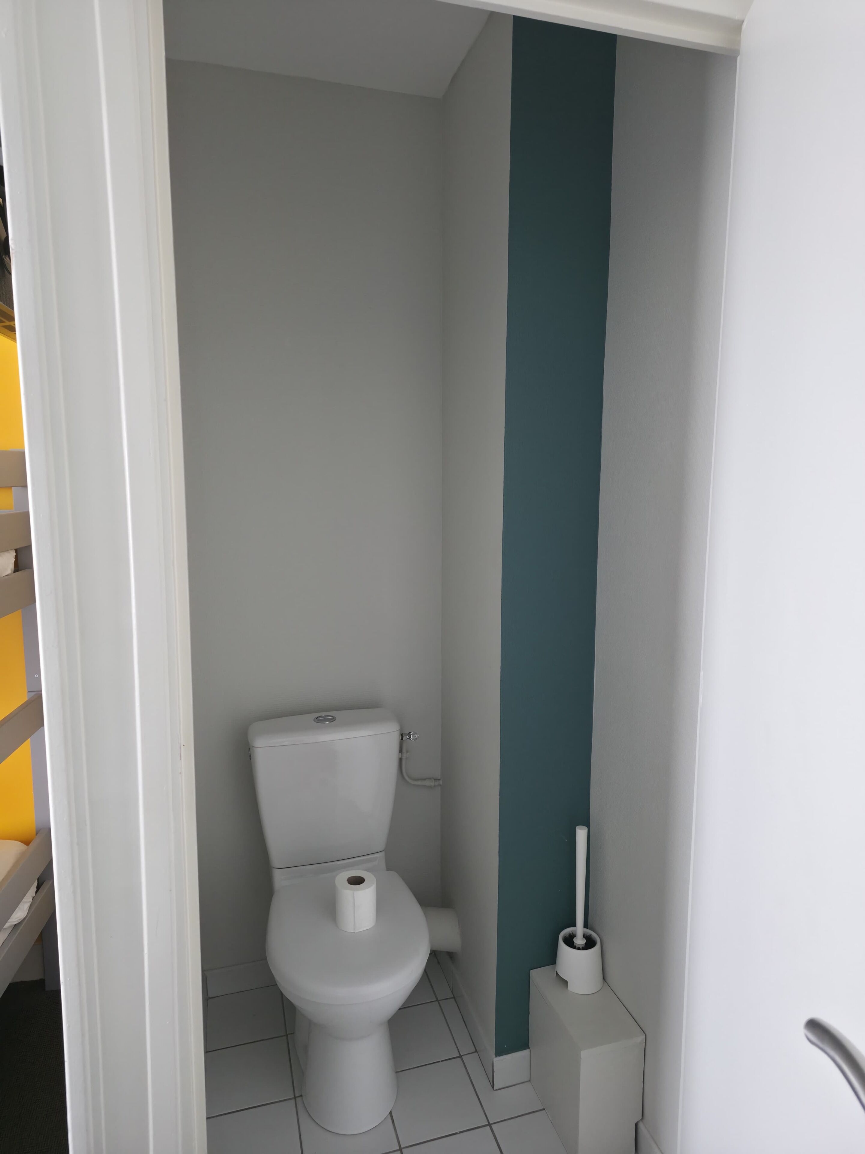 De petites toilettes blanches, avec un rouleau de papier toilette, une brosse de toilette blanche et un mur d’accent de couleur turquoise dans une salle de bain.