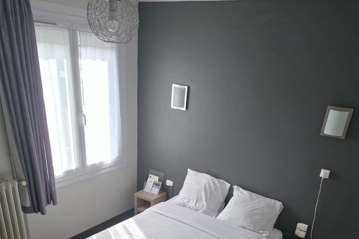 A hotel room with a large bed, a bedside table with a lamp, a window with curtains, and a radiator.