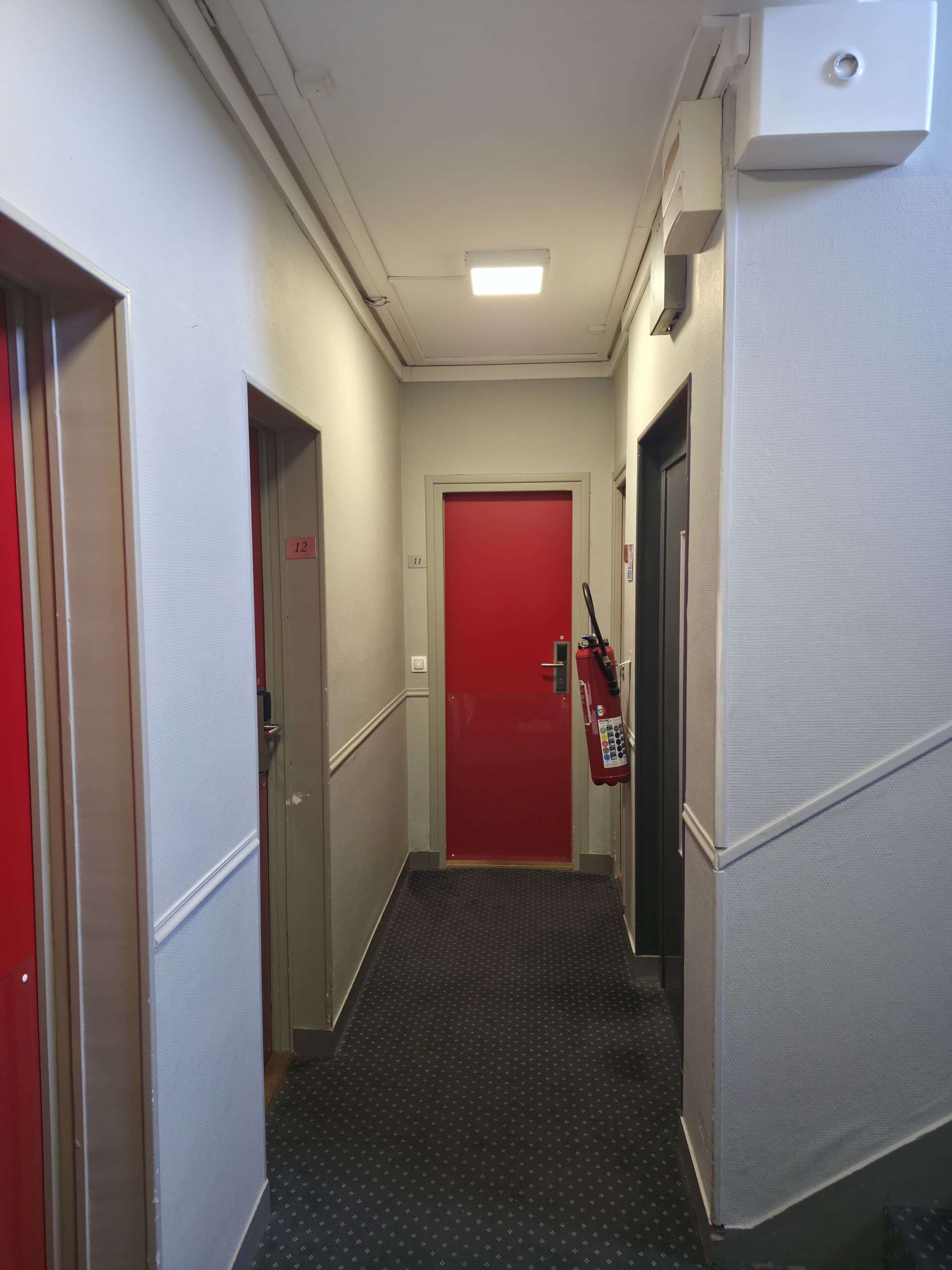 Un pasillo estrecho con una puerta roja al final, un extintor montado en la pared y un panel de control a la derecha.