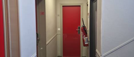 Un pasillo estrecho con una puerta roja al final, un extintor montado en la pared y un panel de control a la derecha.