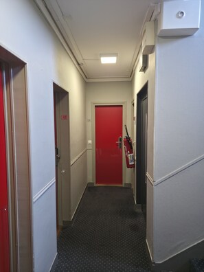 A narrow hallway with a red door at the end, a fire extinguisher mounted on the wall, and a control panel on the right. - The Originals City, Hôtel Le Savoy, Caen (Caen)