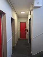 Un pasillo estrecho con una puerta roja al final, un extintor montado en la pared y un panel de control a la derecha.