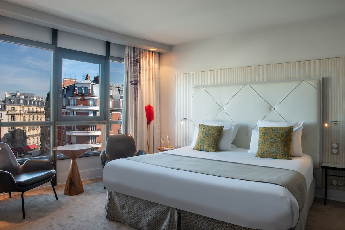 A hotel room with a large bed, a small table, a chair, and a view of the city through large windows.