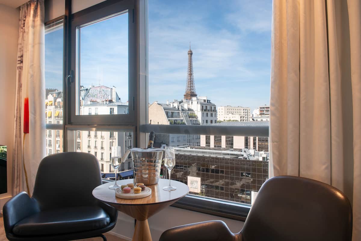 A room with a view of the Eiffel Tower, a table with a bottle and glasses, and two chairs.