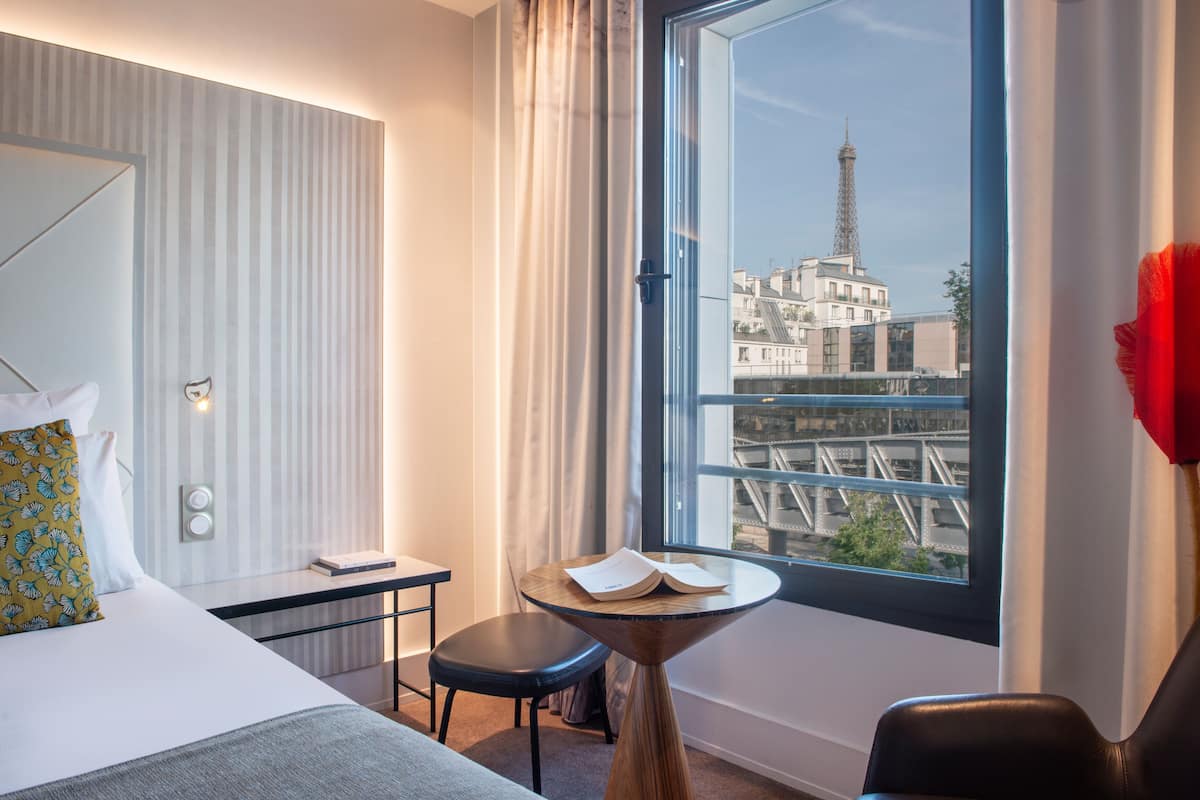 A hotel room with a bed, a small table with a book, a chair, a window with a view of a tower and buildings.