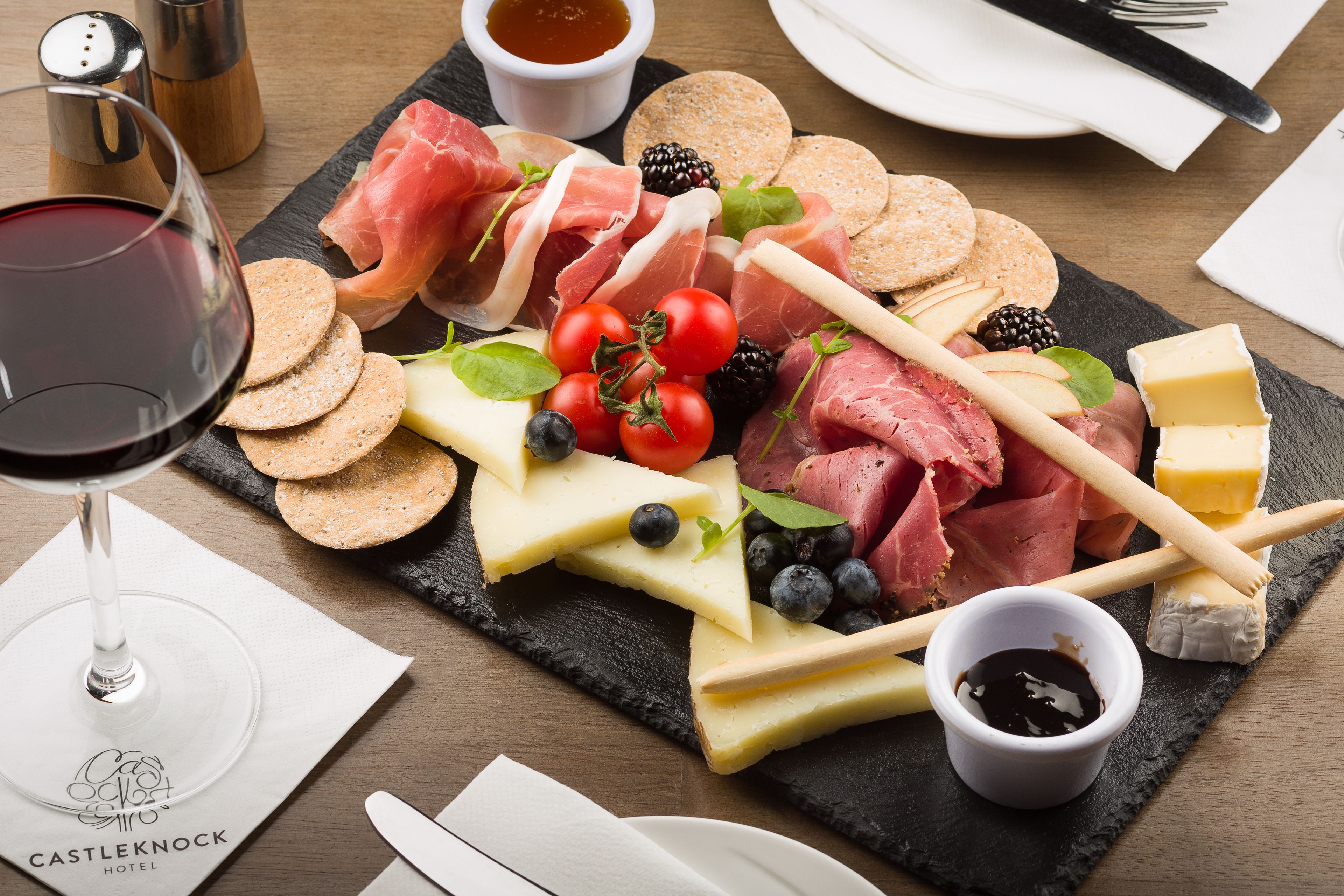 A charcuterie board with various meats, cheeses, and fruits, accompanied by a glass of red wine and a napkin with the hotel's logo.