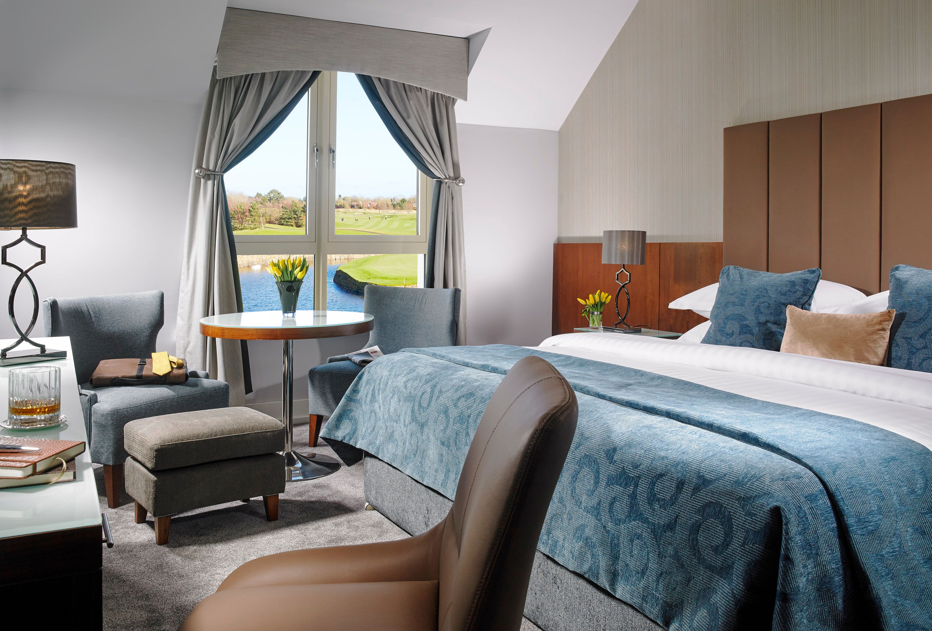 A hotel room with a large bed, a desk with a lamp, a chair, a small table, and a view of a golf course through the window.