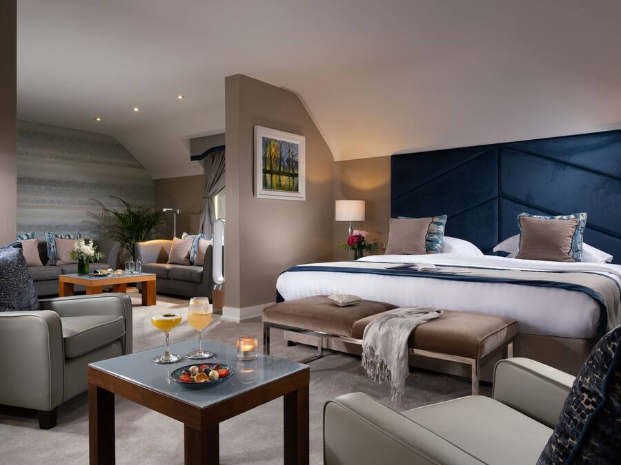 A modern hotel room with a large bed, a sofa, a coffee table with drinks, and a painting on the wall.