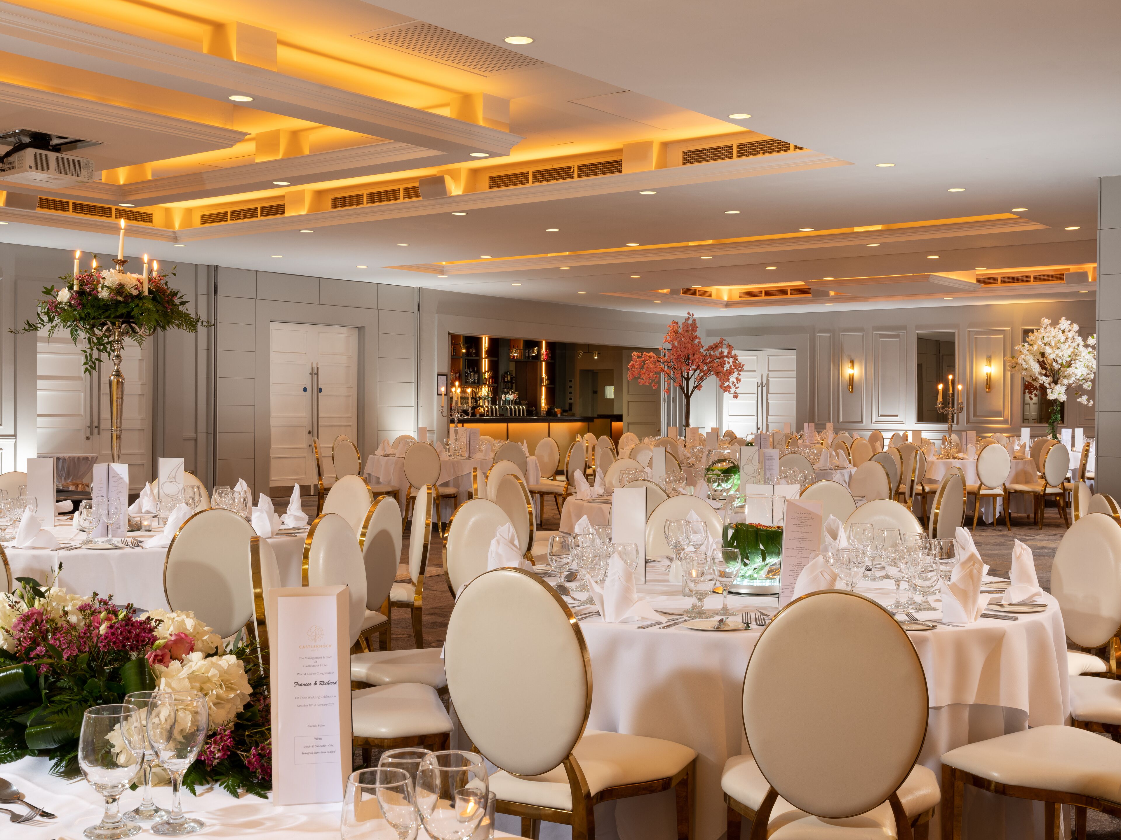 Un salón de banquetes con mesas redondas dispuestas para un evento formal, con manteles blancos, sillas doradas y centros de mesa florales.