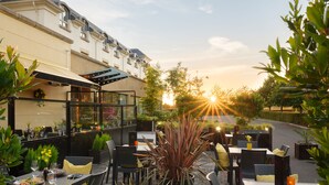 Un área de comedor al aire libre con mesas y sillas, rodeada de plantas y un edificio con terraza.