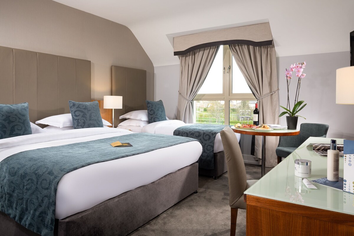 A hotel room with two beds, a desk, a chair, a window with curtains, a potted plant, and a table with a bottle and glasses.