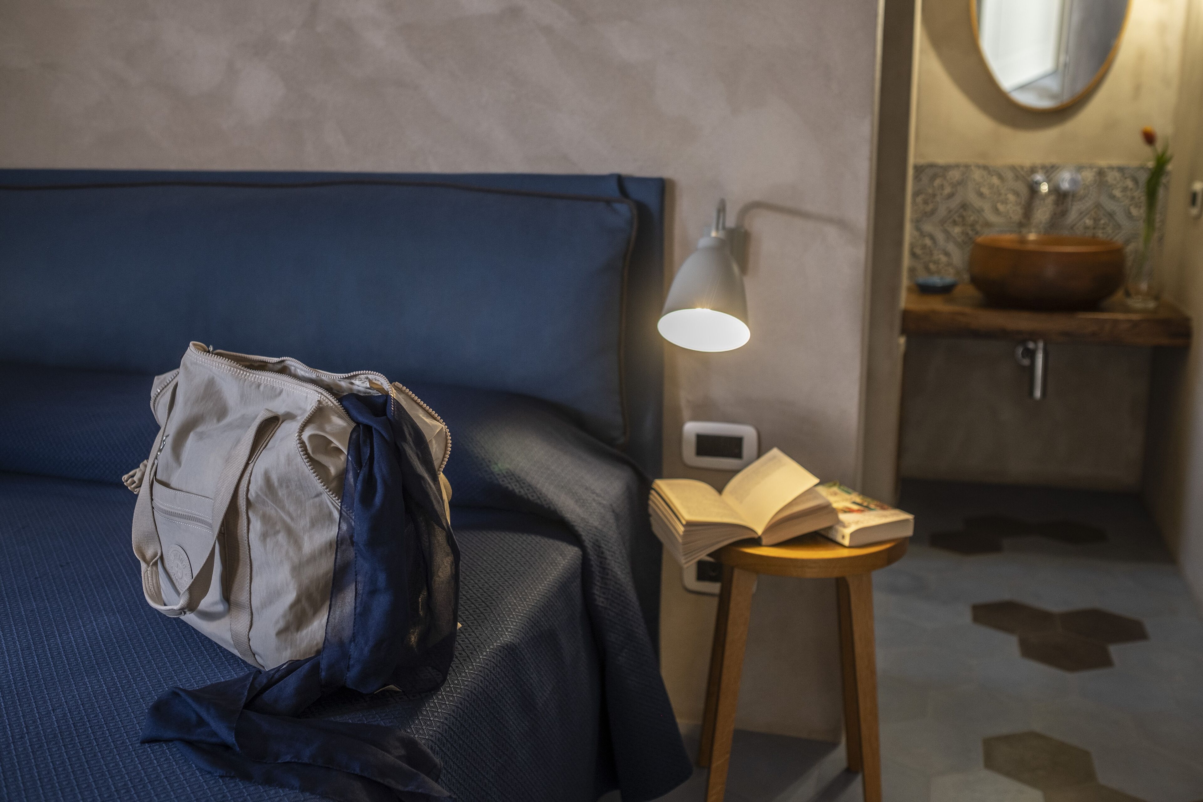 Un lit bien fait, avec une tête de lit bleue, une lampe de chevet, une petite table de nuit en bois, et une salle de bain visible au fond.