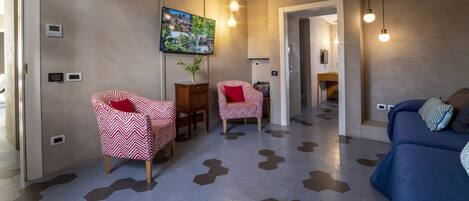 A modern living room with a blue sofa, two red and white patterned chairs, a dining table with chairs, a painting on the wall, and hexagonal patterned flooring.