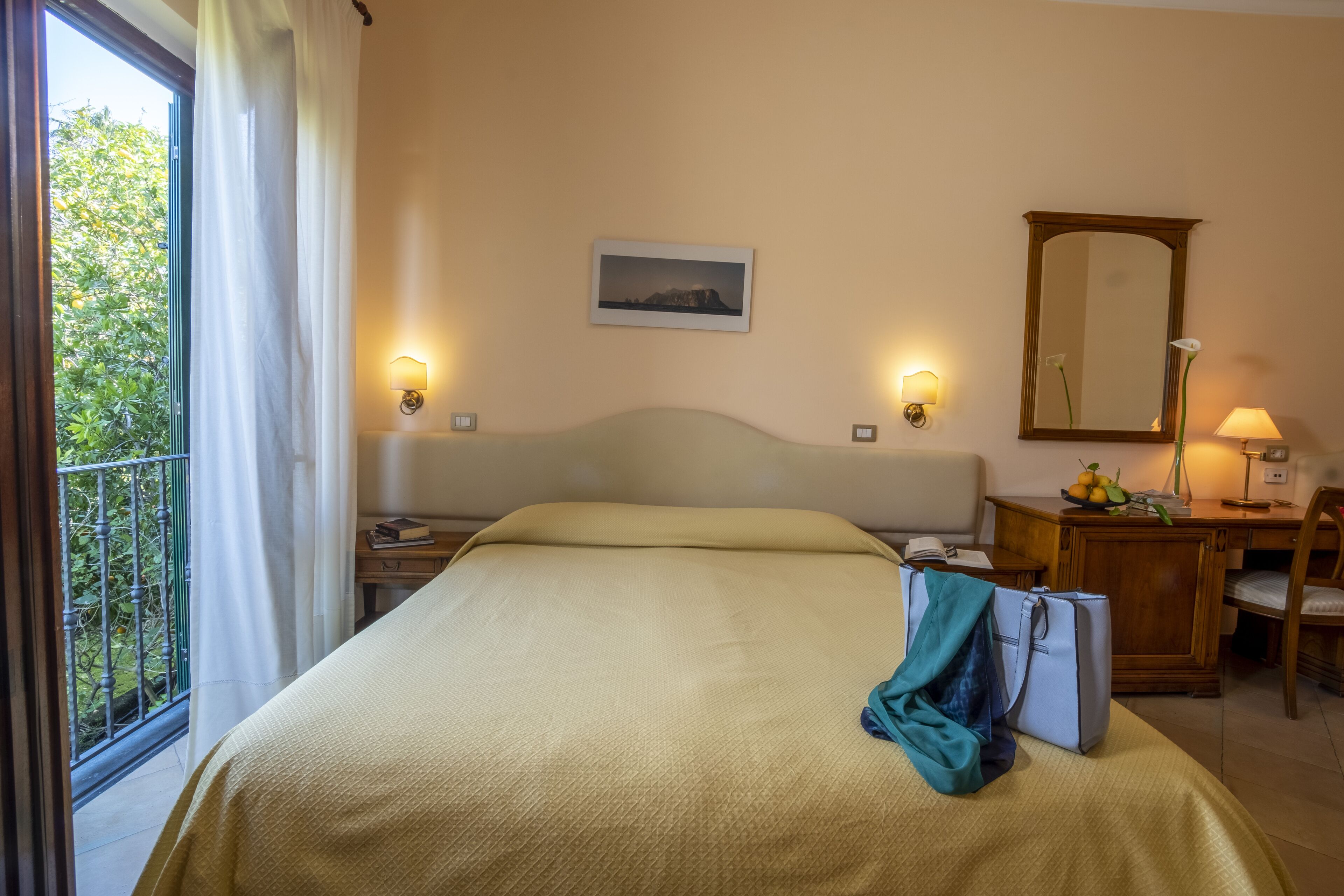Una camera d'albergo con un letto, una scrivania, una sedia, una lampada, uno specchio e una finestra con tende.