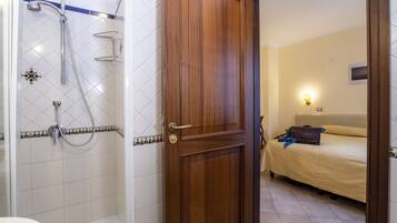 Ein Badezimmer mit Dusche, Toilette und einer Holztür, die in einen Schlafbereich mit Bett und Nachttisch führt.