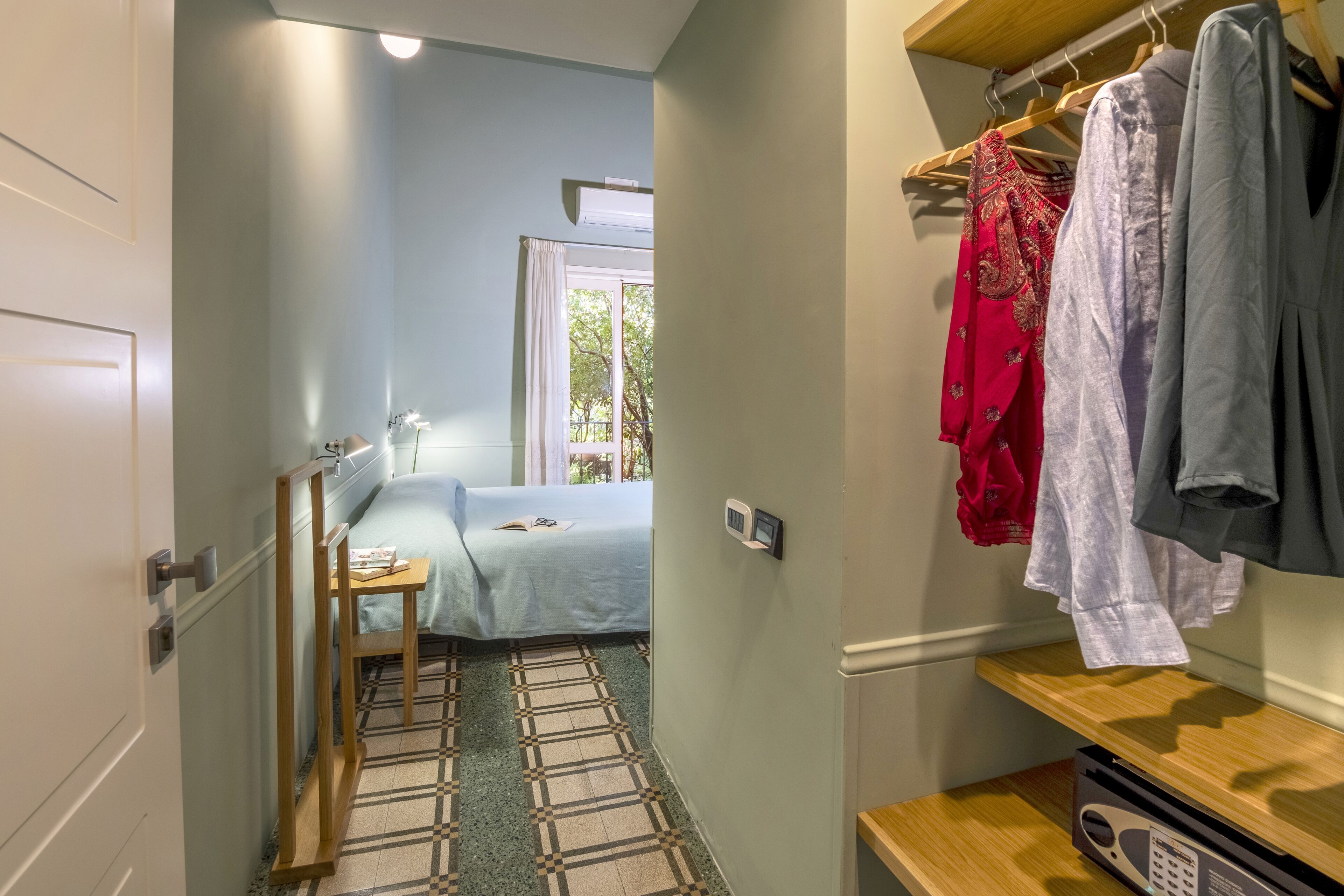 A hotel room with a bed, a desk, a chair, a window with a view, and a clothing rack with various garments.