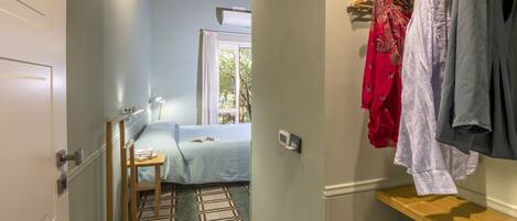 A hotel room with a bed, a desk, a chair, a window with a view, and a clothing rack with various garments.
