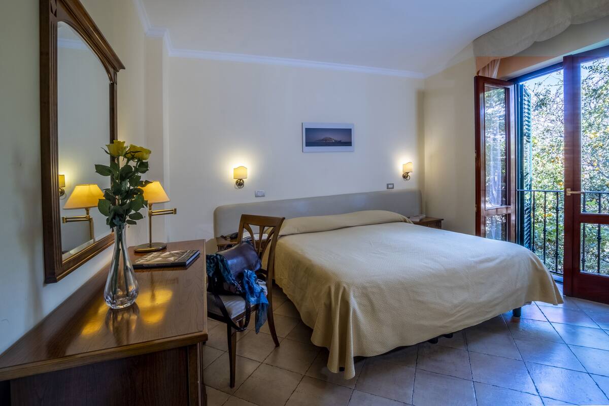 Un dormitorio con cama, mesitas de noche, un espejo, un jarrón con flores y vistas a la vegetación a través de una ventana.