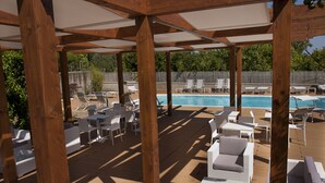 Un espacio al aire libre techado con vigas de madera, sillones blancos y una piscina.
