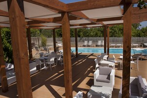 Un espacio al aire libre techado con vigas de madera, sillones blancos y una piscina.