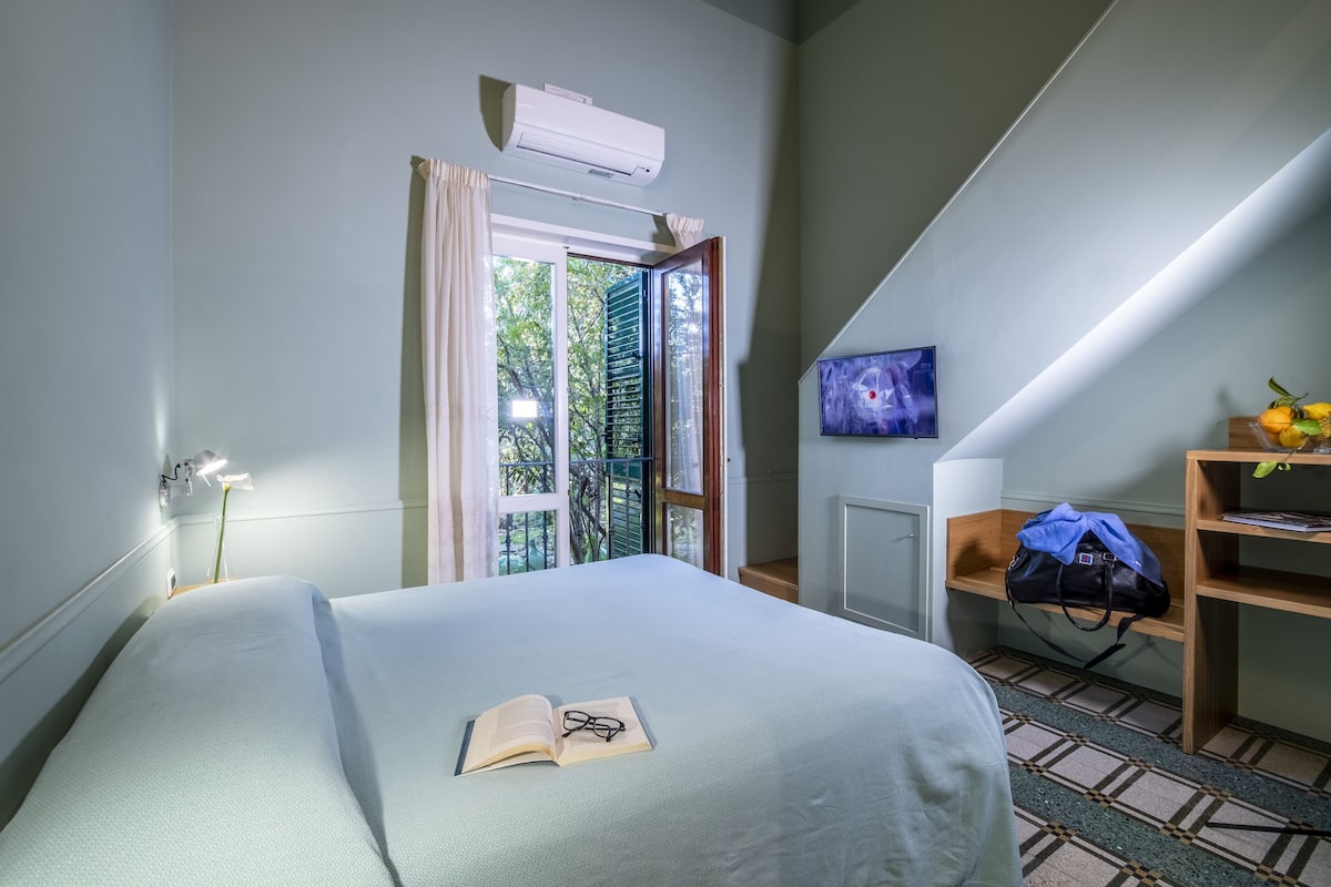 Un dormitorio con cama, ventana abierta, escritorio con bolso y aire acondicionado de pared.