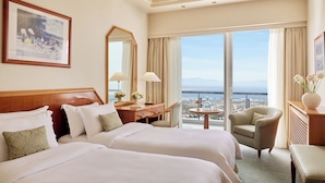 A hotel room with two beds, a desk, a chair, and a view of the city. - Hotel Panorama (Pylaia-Chortiatis)