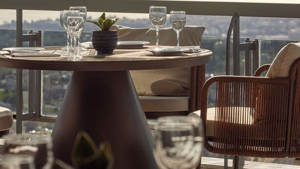 A dining table with chairs and wine glasses, overlooking a body of water and distant mountains. - Hotel Panorama (Pylaia-Chortiatis)
