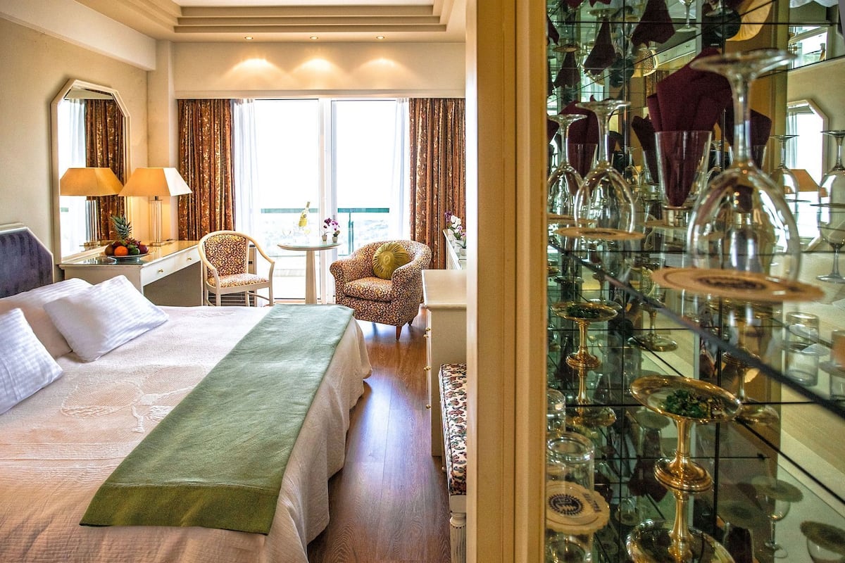 A hotel room with a bed, two chairs, a table with fruit, and a glass display of cutlery.