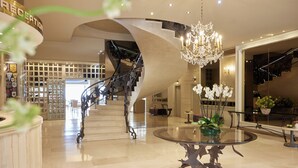 A grand lobby with a spiral staircase, a glass table with a metal base, and a chandelier. - Hotel Panorama (Pylaia-Chortiatis)