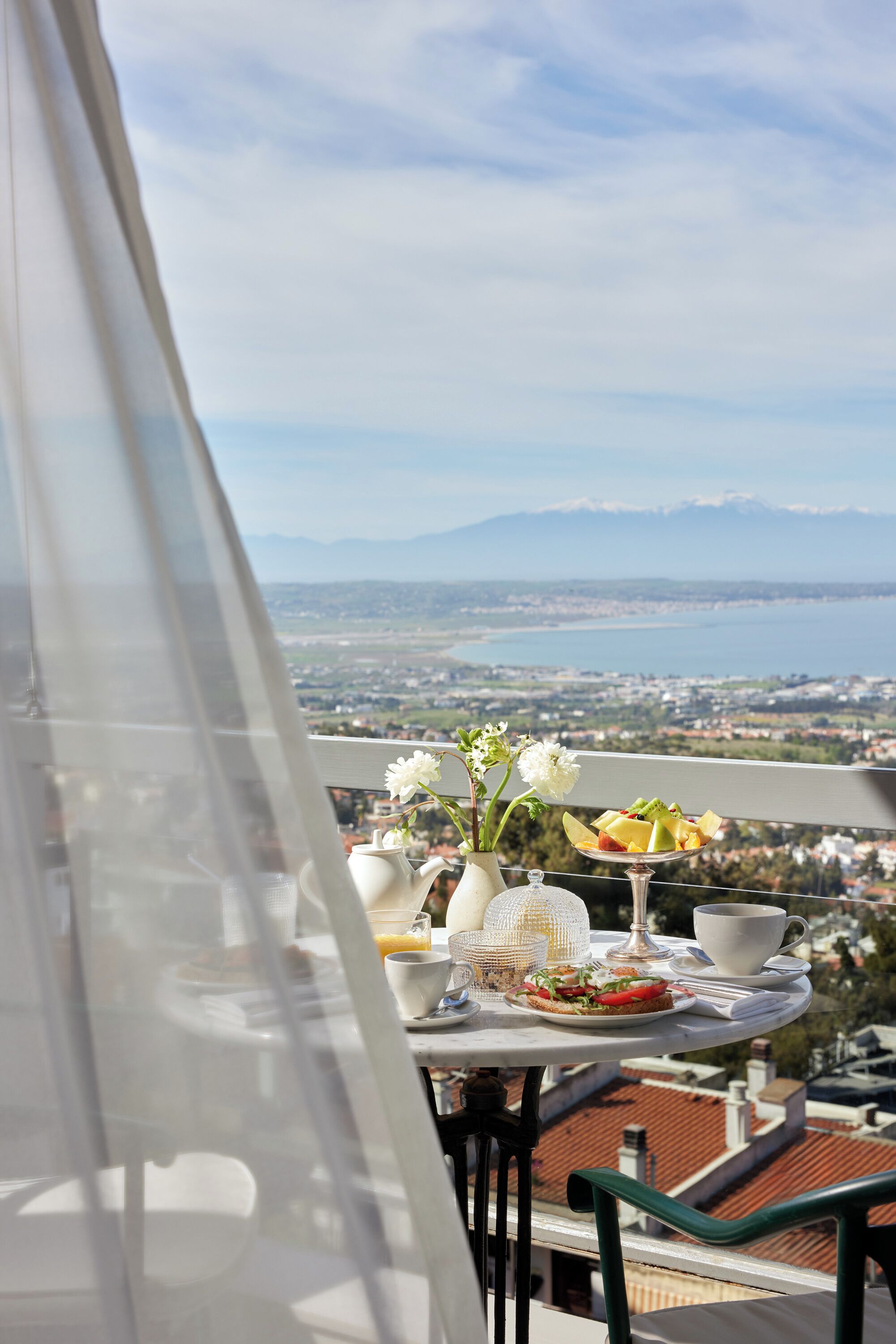 Un balcón con una mesa lista para el desayuno, un jarrón con flores y una vista panorámica de un lago y montañas.