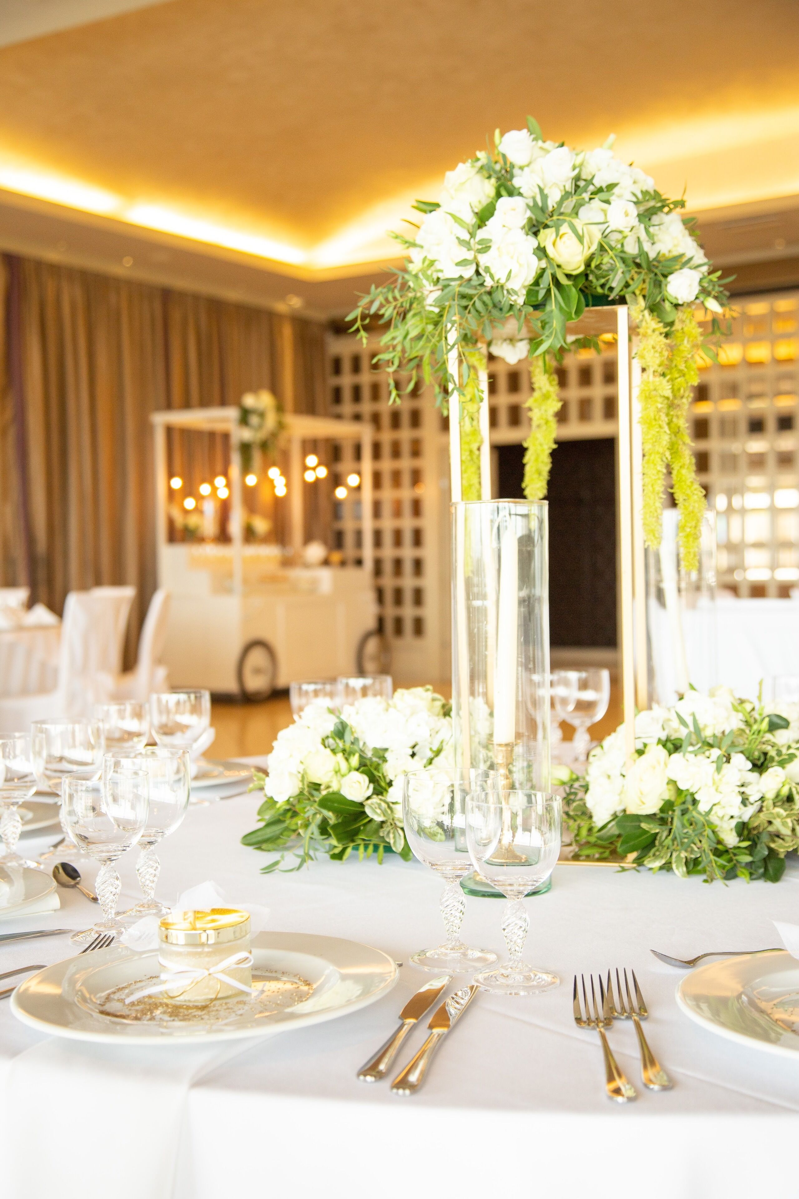 Mesa dispuesta con platos blancos, cubiertos y copas de cristal, decorada con flores y follaje blanco en una habitación luminosa con ambiente cálido.