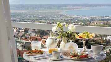 Ett balkongbord med frukostmat och en vas med blommor, med utsikt över en stad och berg.