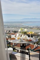 Mesa en balcón con alimentos para el desayuno y un jarrón de flores, con vista a un paisaje urbano y montañas.