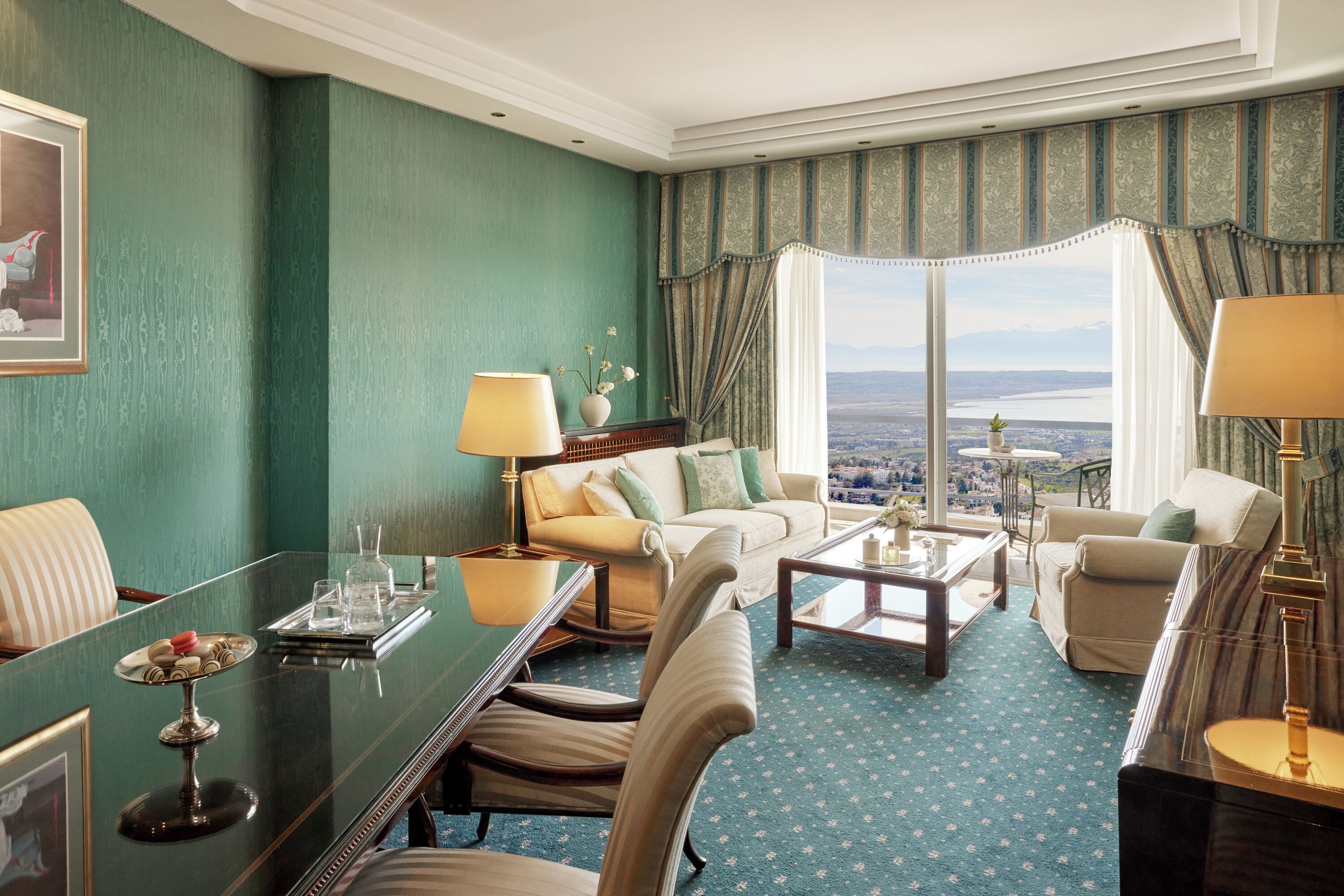 A hotel room with a large window offering a city view, a seating area with a sofa and chairs, and a dining table with a tray of glasses and a vase of flowers.