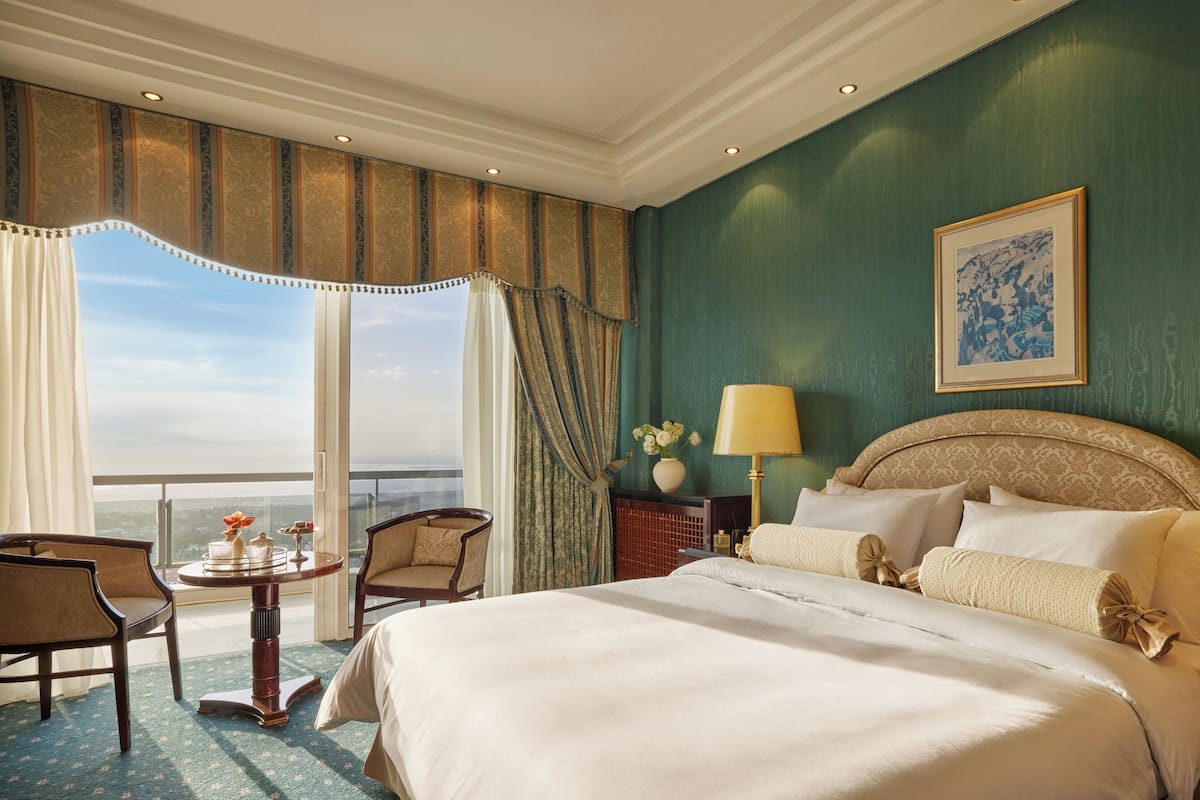 A hotel room with a large bed, two bedside lamps, a chair, a small table with a vase of flowers, and a view of the ocean through large windows.