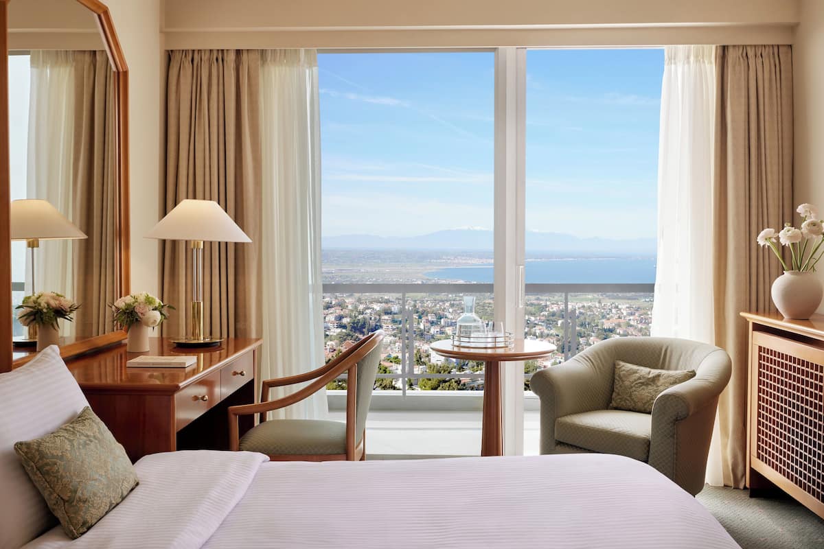 A hotel room with a large bed, two chairs, a desk, and a view of a cityscape through large windows.