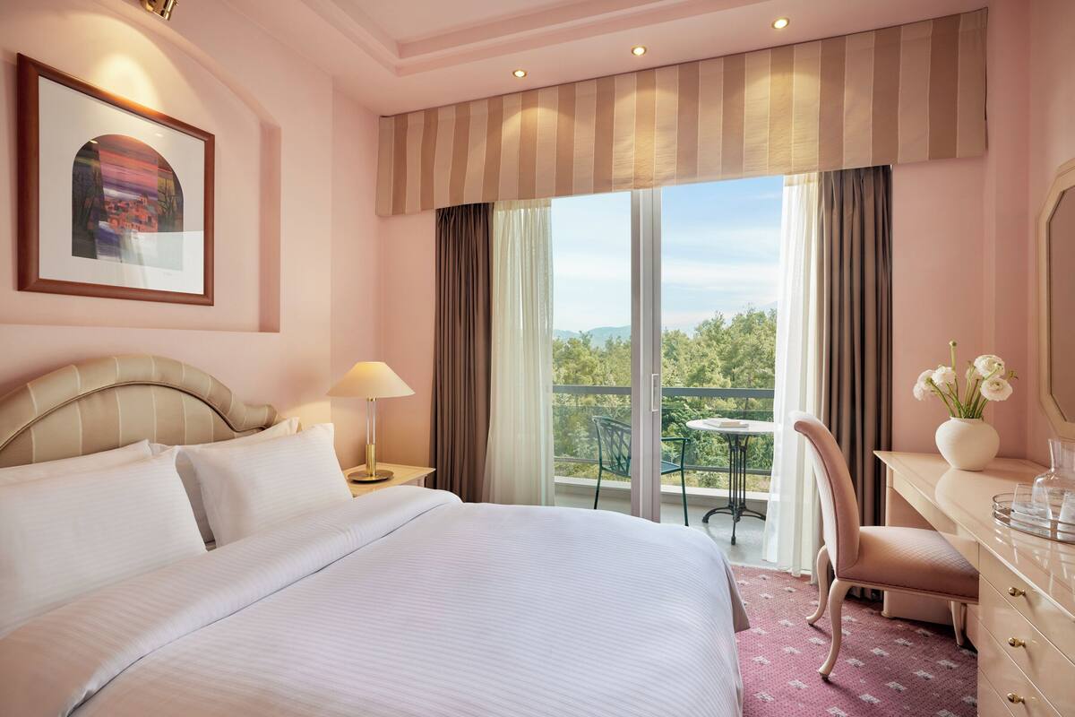 A hotel room with a large bed, a desk with a chair, a window with curtains, a framed picture on the wall, and a balcony with a table and chairs.