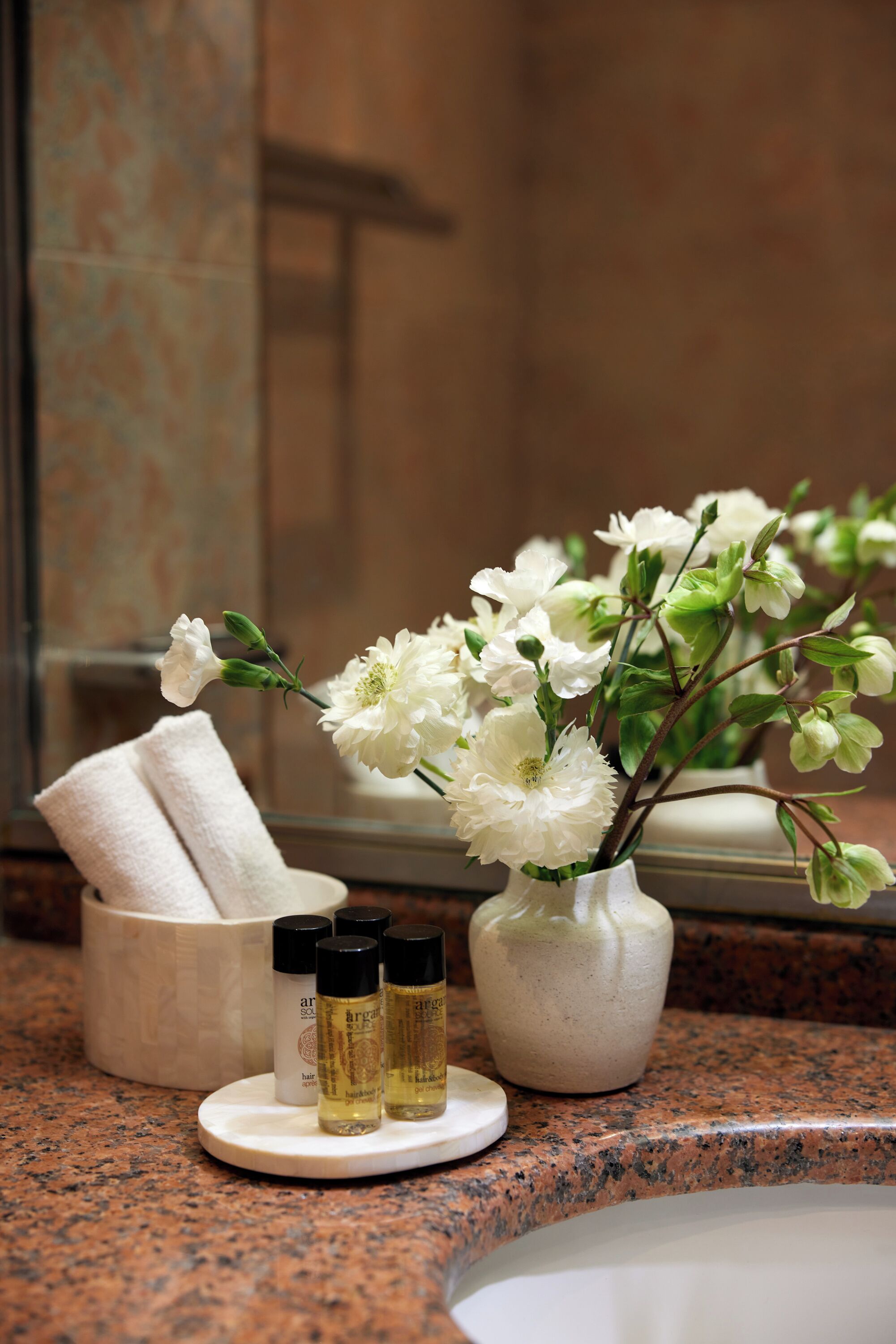 Un tocador de baño con un florero de flores blancas, una toalla y tres botellas de productos para el cabello.