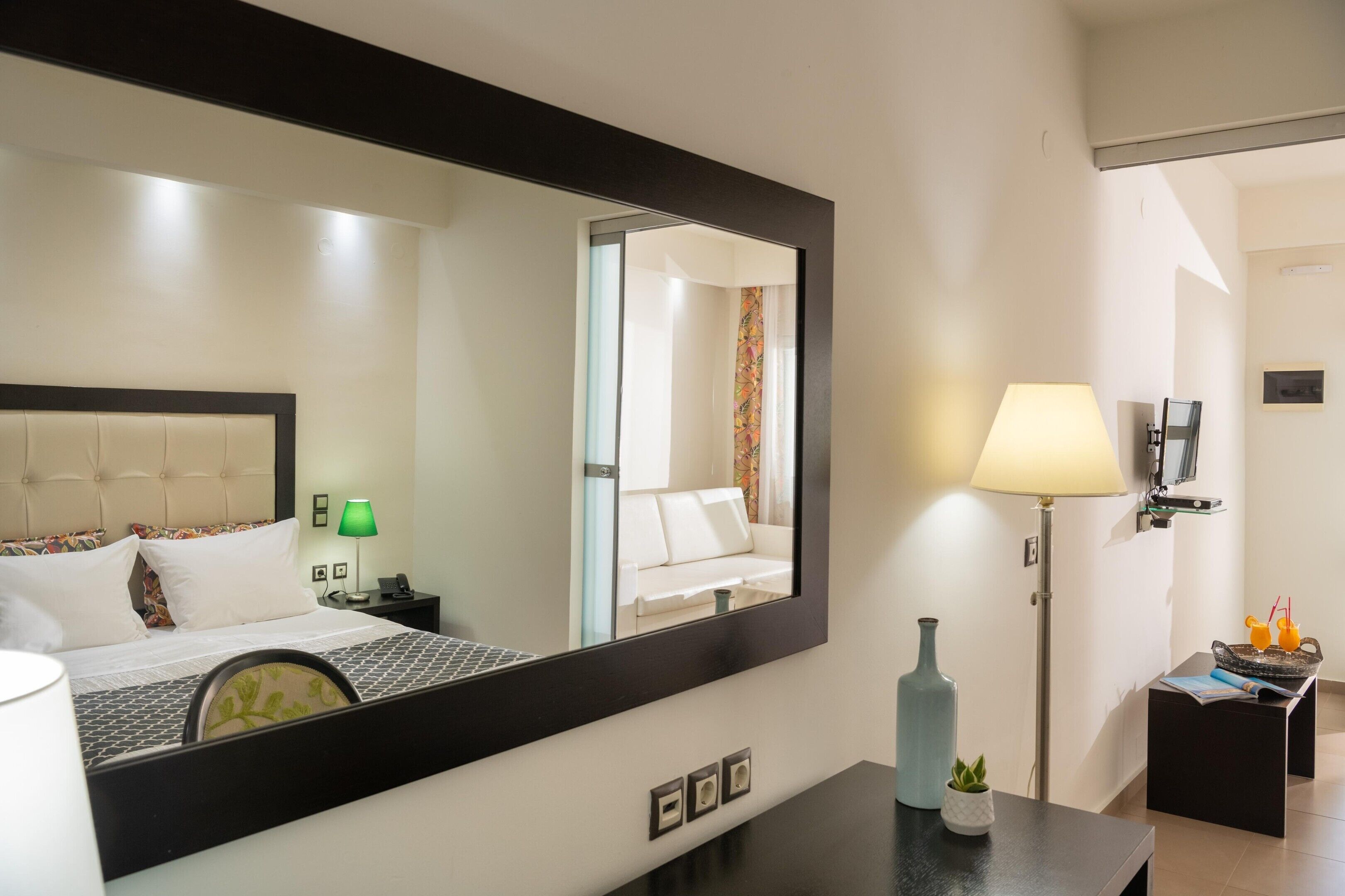 A hotel room with a bed, a large mirror, a nightstand, a lamp, and a small table with a vase and a plant.
