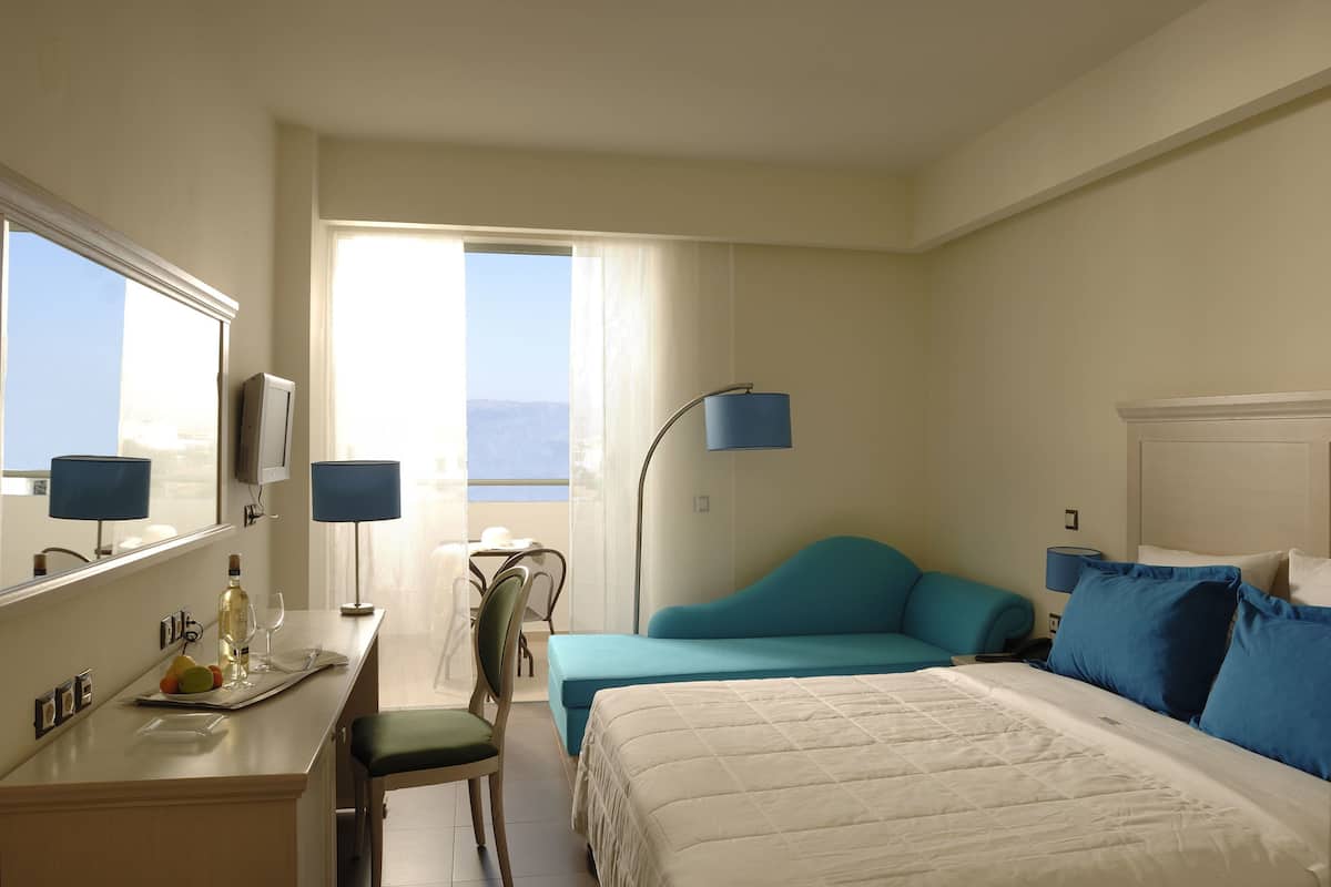 Ein Hotelzimmer mit Bett, Schreibtisch mit Stuhl, einem kleinen Tisch ebenfalls mit Stuhl, einer Lampe und Blick ins Freie.