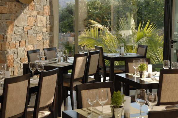 Un restaurante con paredes de piedra, grandes ventanales y plantas tropicales en el exterior. Las mesas están dispuestas con copas de vino y platos.