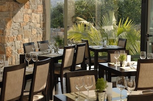A restaurant with stone walls, large windows, and outdoor tropical plants. Tables are set with wine glasses and plates.