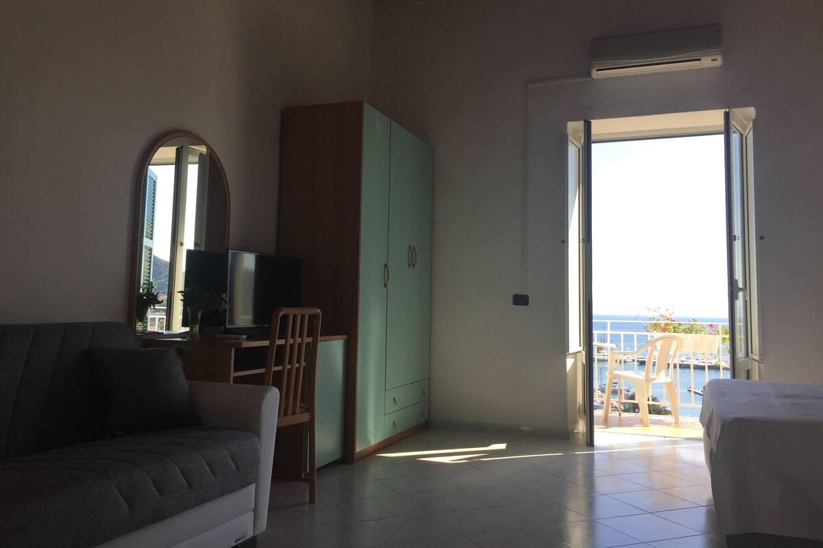 A living room with a sofa, TV, and a cabinet. There's a door leading to a balcony with a view of the sea.