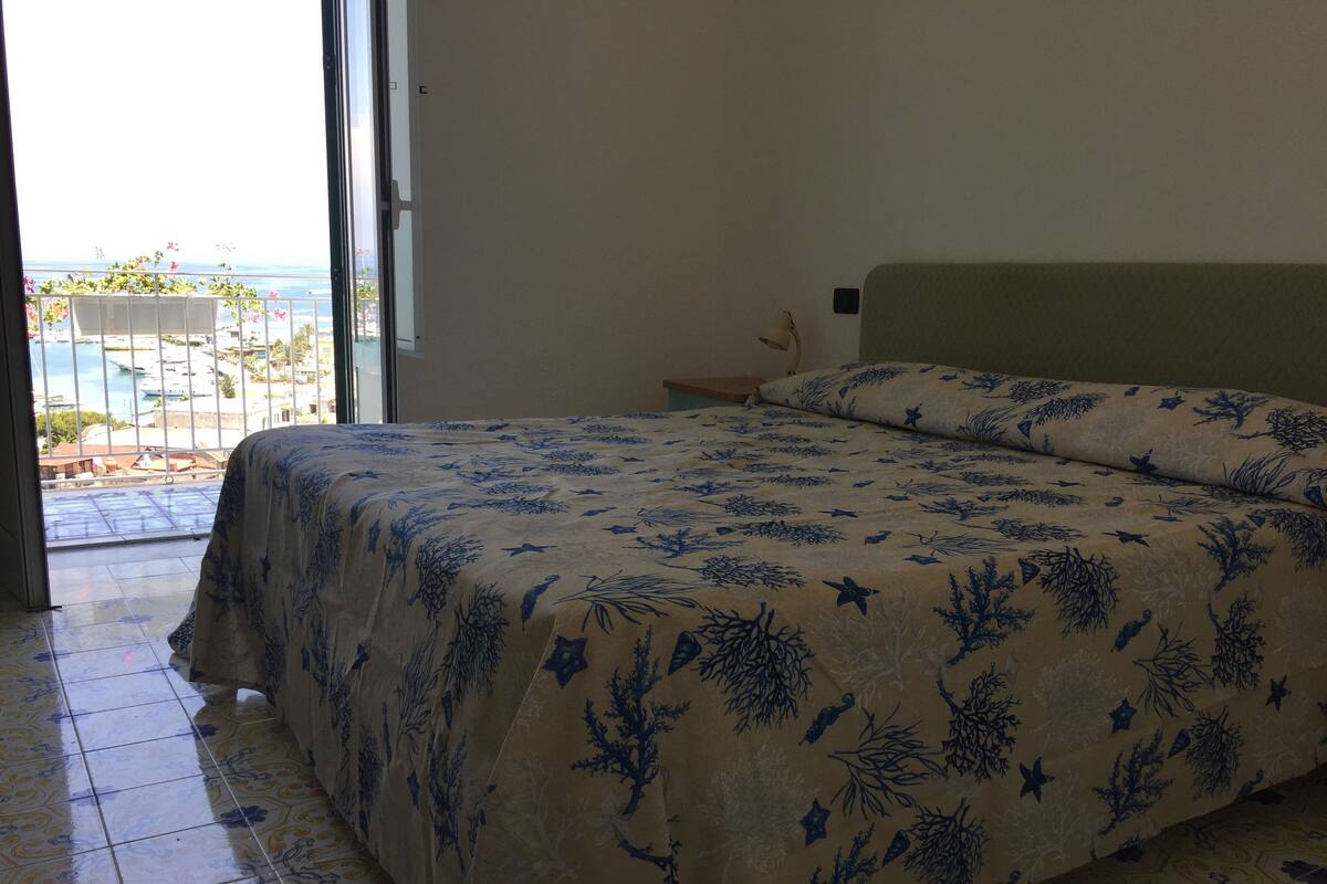 A bedroom with a bed, a nightstand, and a view of the sea through a large window.