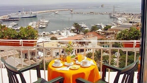 Un balcón con una mesa lista para el desayuno, con vistas a un puerto deportivo con barcos y un pueblo costero.