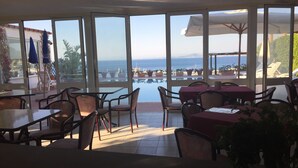 Un comedor interior con amplios ventanales que dan a una piscina y al mar. Mesas y sillas dispuestas para comer.