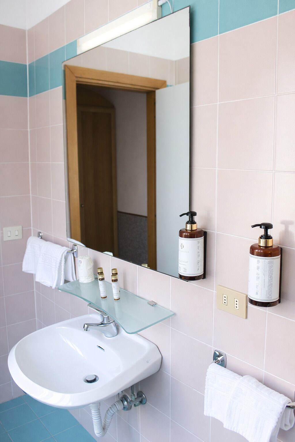 Un baño con un lavamanos blanco, una repisa de vidrio con artículos de aseo, un espejo y un perchero para toallas.