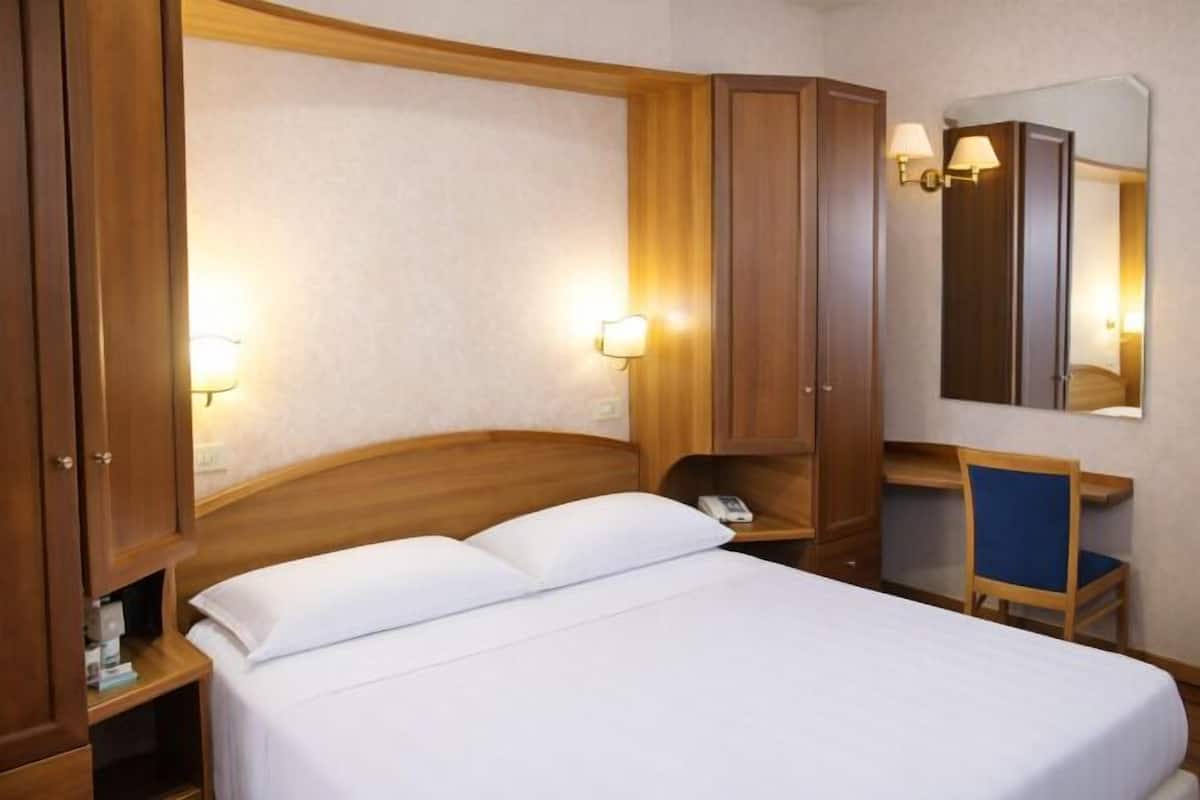 A hotel room with a bed, wooden wardrobe, bedside table, lamp, and a mirror.